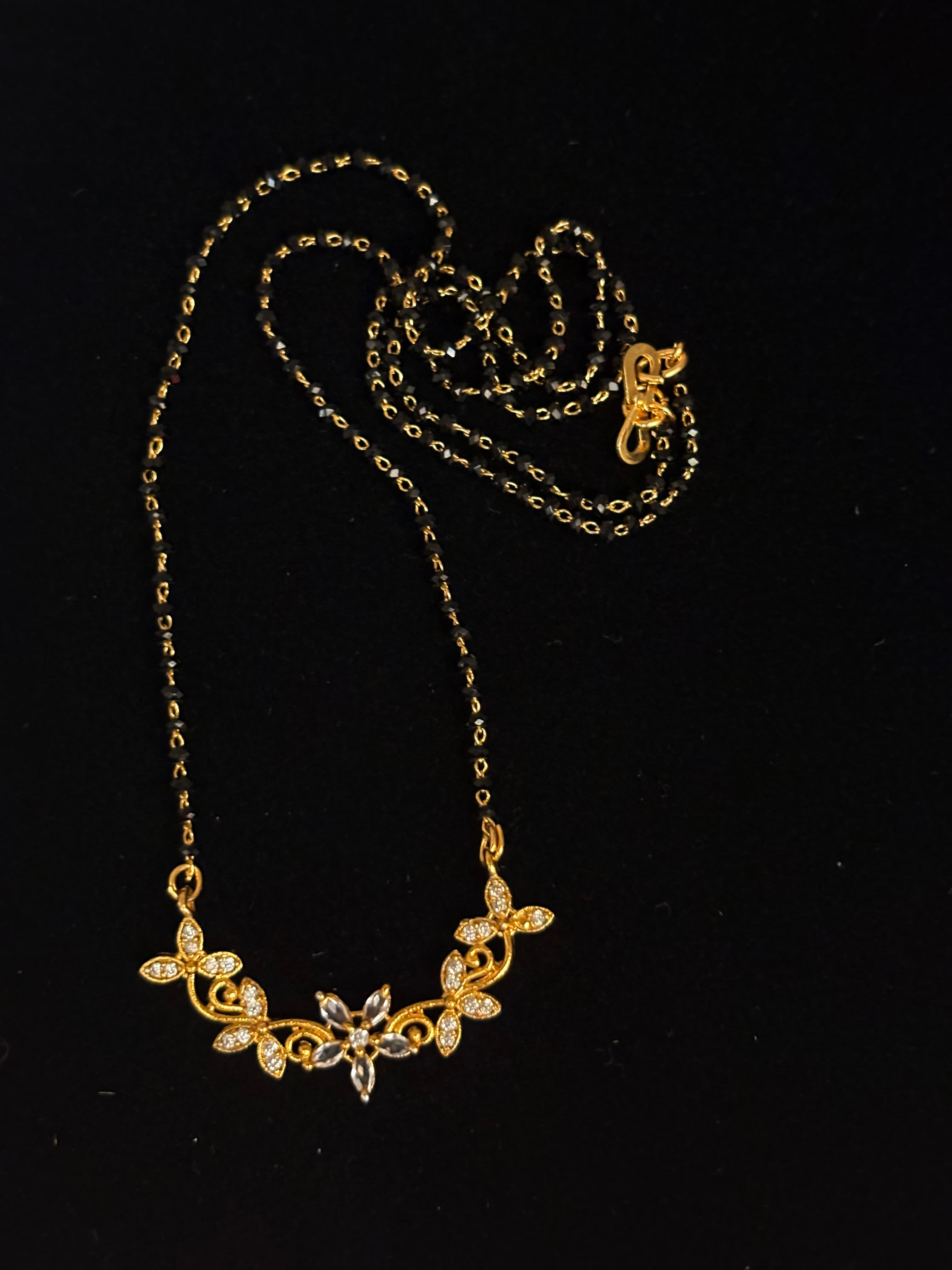 Mangalsutra with black bead chain and gold-toned floral pendant with clear accents on black background