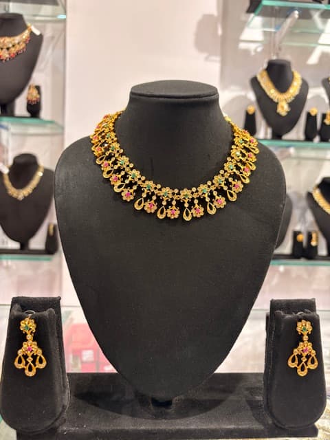 Gold-tone choker necklace with floral motifs, green and pink accents and matching teardrop earrings on black display